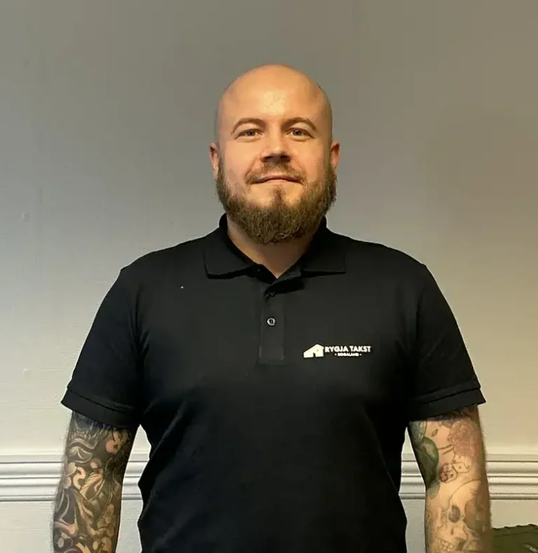 Bildet viser en takstmann fra Rygja Takst som står foran en nøytral vegg, iført svart polo med logo, klar til å utføre takst- og verdivurderingstjenester.