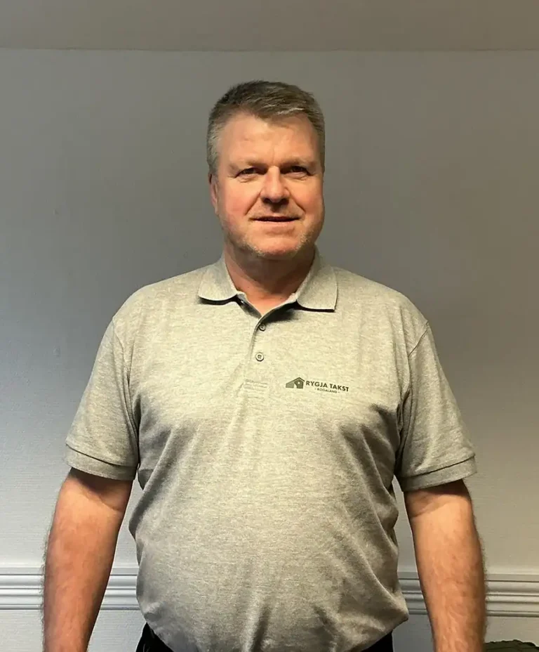 Bilde av en mann i lys grå polo med Rygja Takst-logo, står foran en enkel vegg, klar for takseringstjenester.